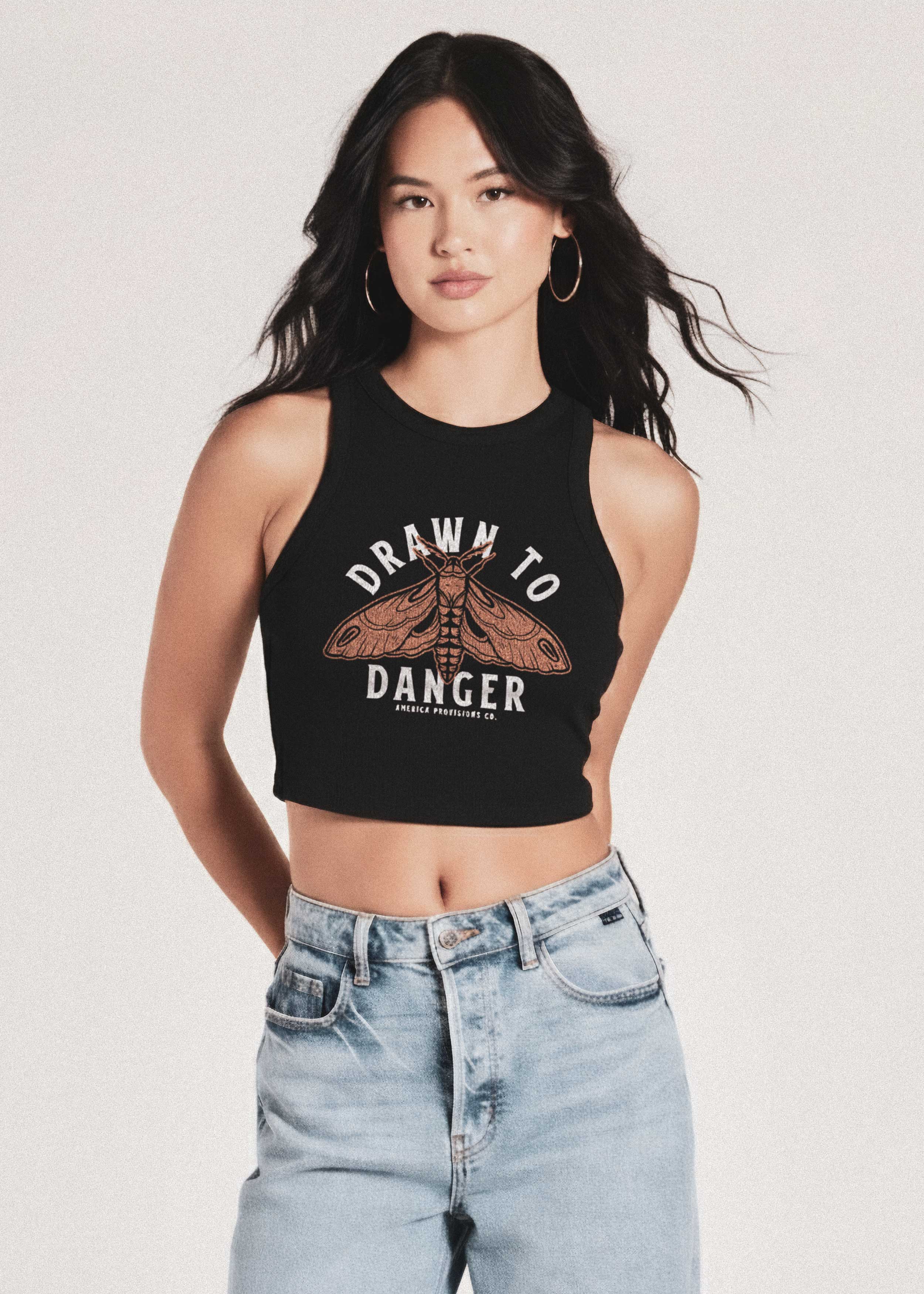 Woman wearing a black tank top with 'Drawn to Danger' graphic and light blue jeans on a white background